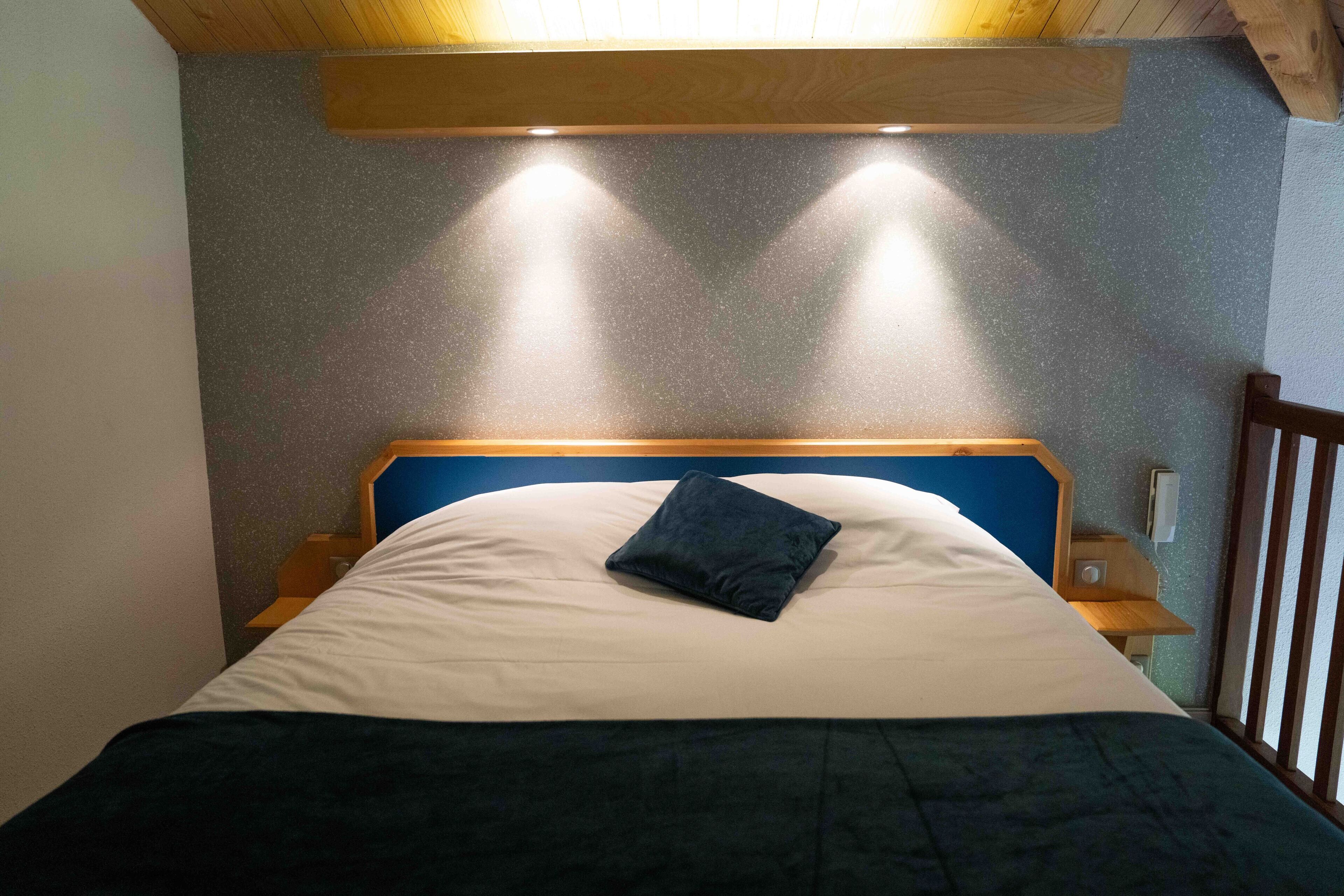 A bed with a blue headboard, a white pillow, and a dark blue blanket. There are two bedside tables with lamps.