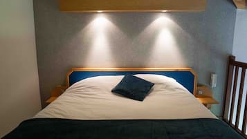 Een bed met een blauw hoofdeinde, een wit kussen en een donkerblauwe deken. Twee nachtkastjes met lampen staan ernaast.