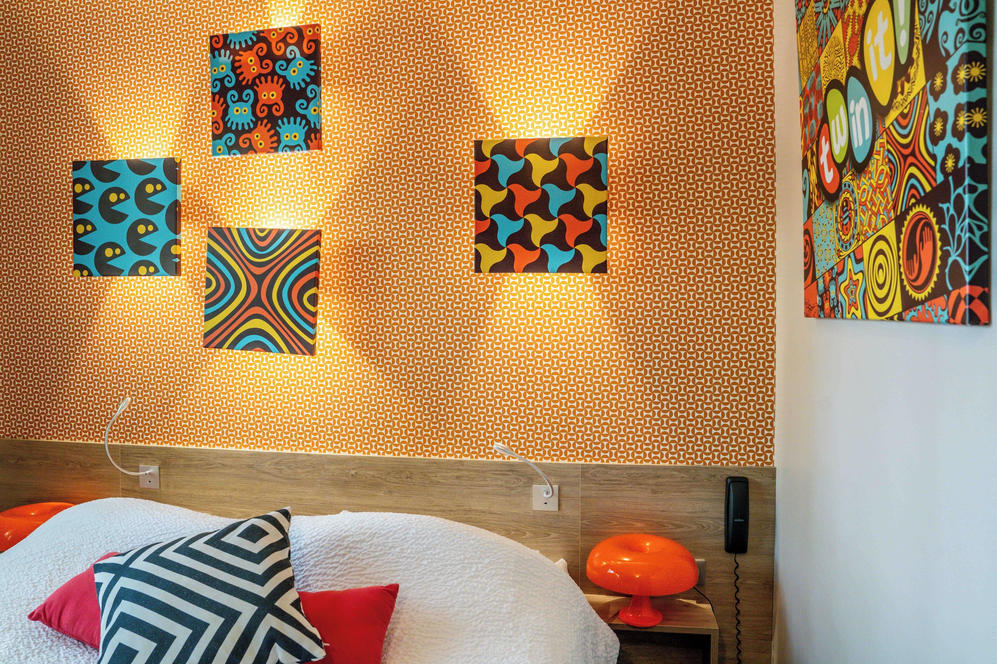 Una habitación de hotel con una pared estampada, una cama con almohadas blancas y rojas, y una mesita de noche con una lámpara.