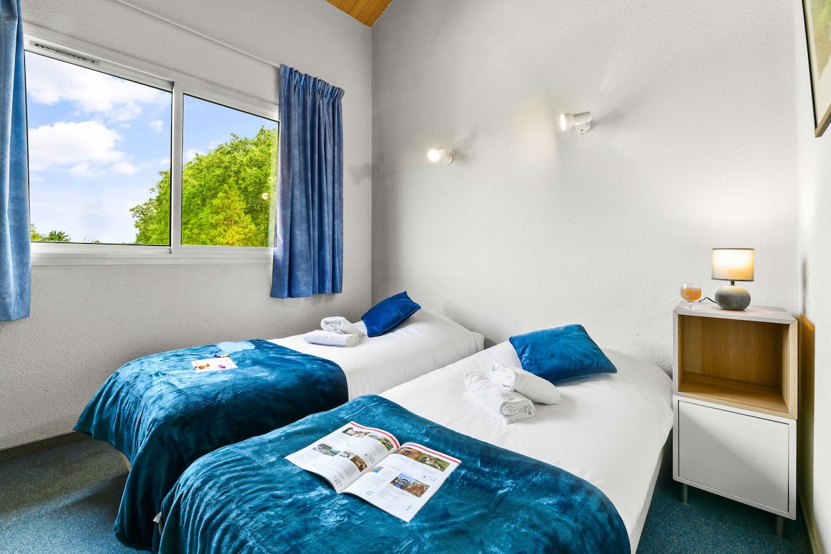 Habitación de hotel con dos camas, una ventana con cortinas, una mesita de noche con lámpara y vistas a zonas verdes.