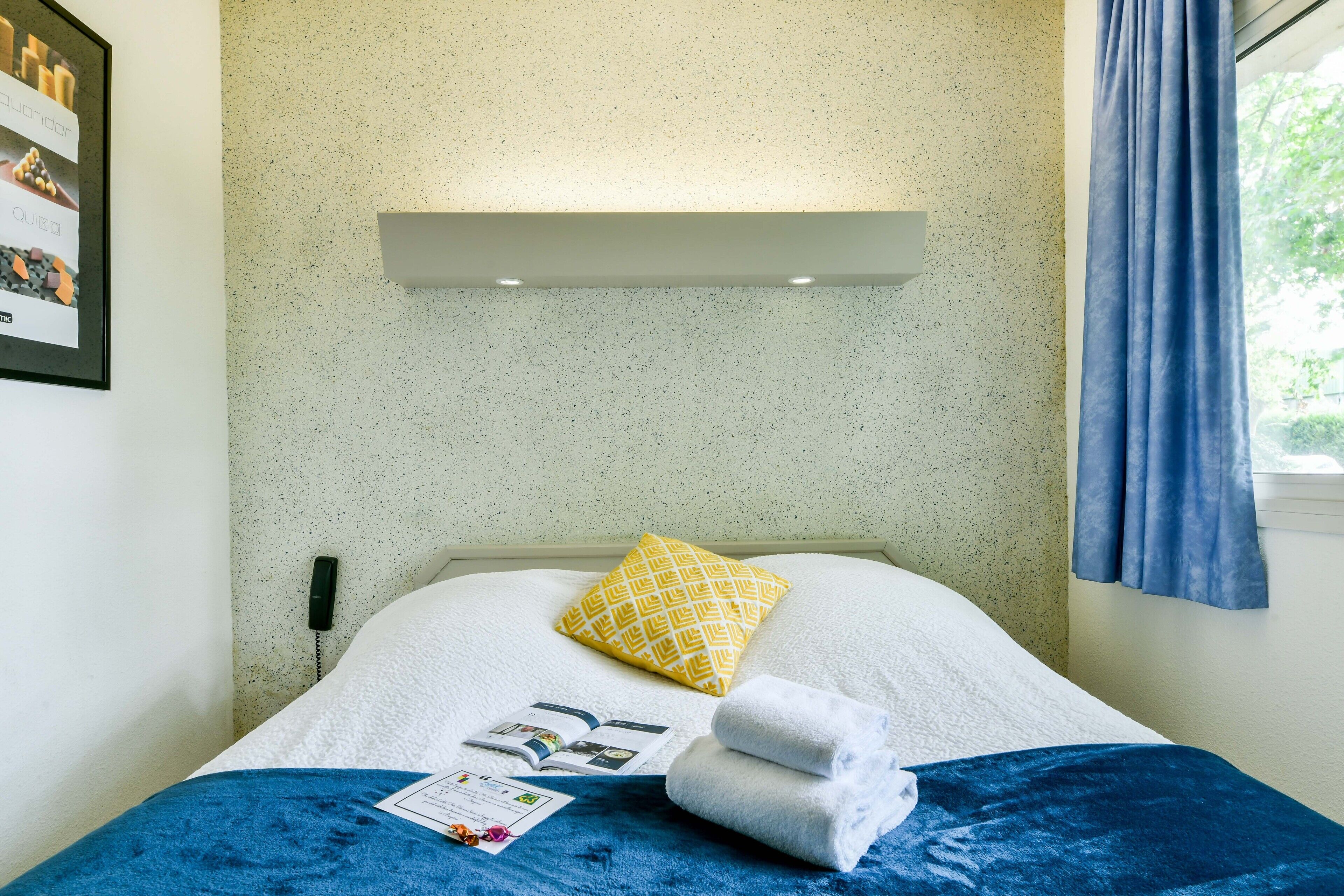 A neatly made bed with a white and blue bedspread, a yellow patterned pillow, and a folded towel.