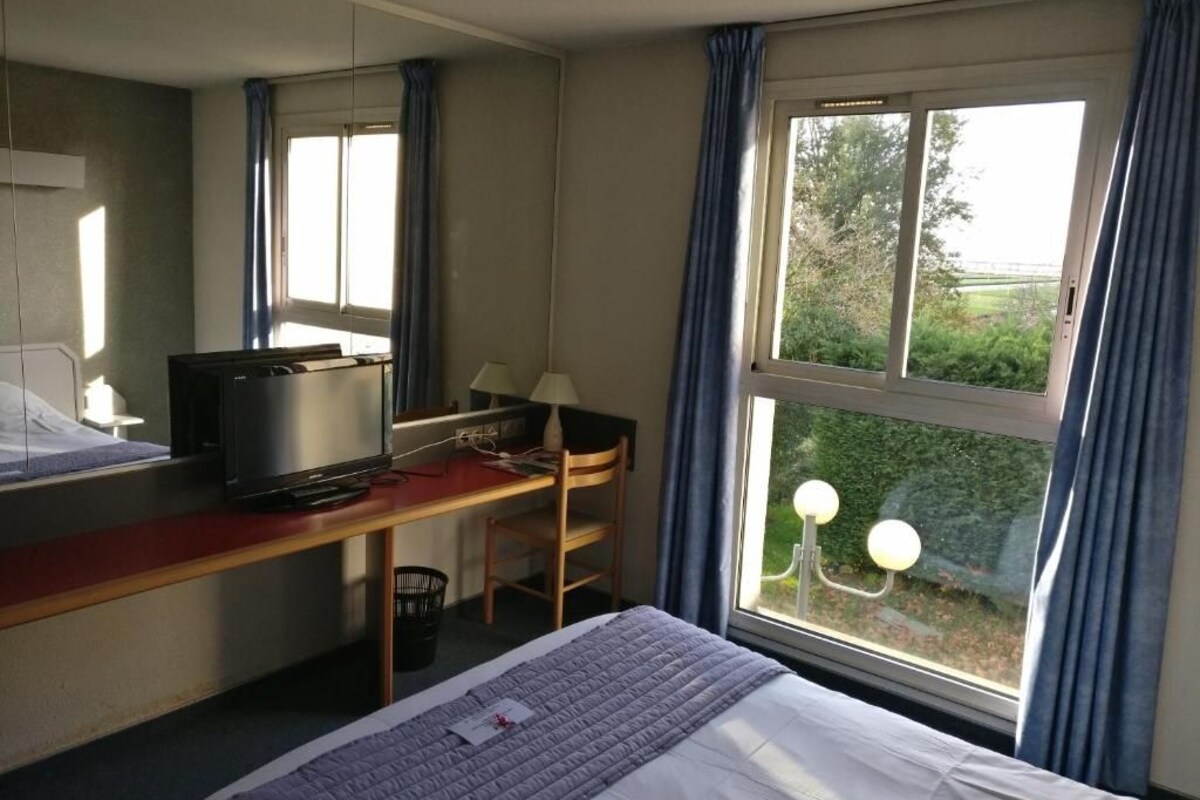 A hotel room with a bed, a desk with a computer and a TV, a chair, and a window with curtains.
