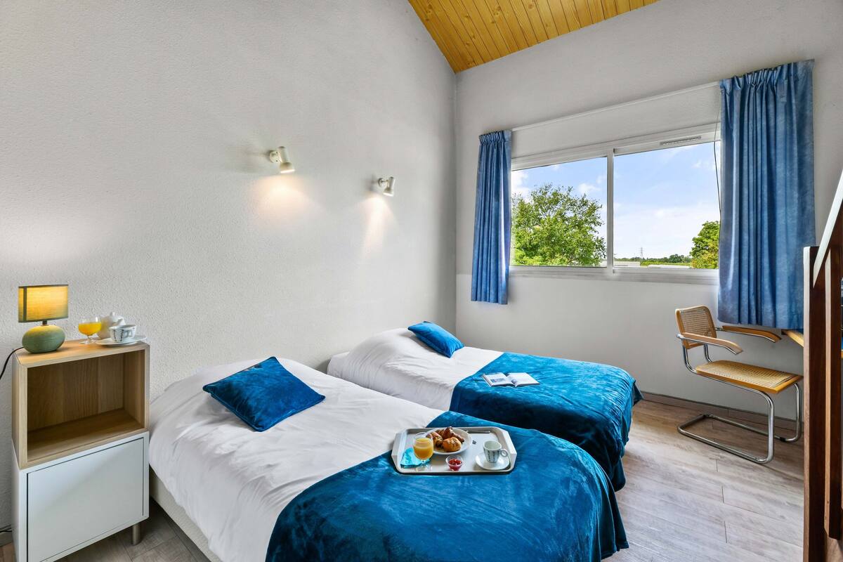 Habitación de hotel con dos camas, una mesita de noche de madera con una lámpara, una bandeja con alimentos para el desayuno y una ventana con cortinas.