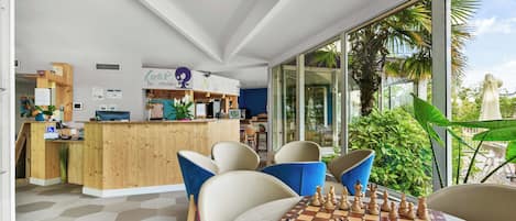 Un intérieur moderne avec un comptoir d’accueil en bois, des chaises bleues, un jeu d’échecs et de grandes fenêtres donnant sur un espace verdoyant.