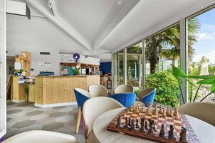 Un interior moderno con un mostrador de recepción de madera, sillas azules, un tablero de ajedrez y grandes ventanales con vistas a la vegetación.