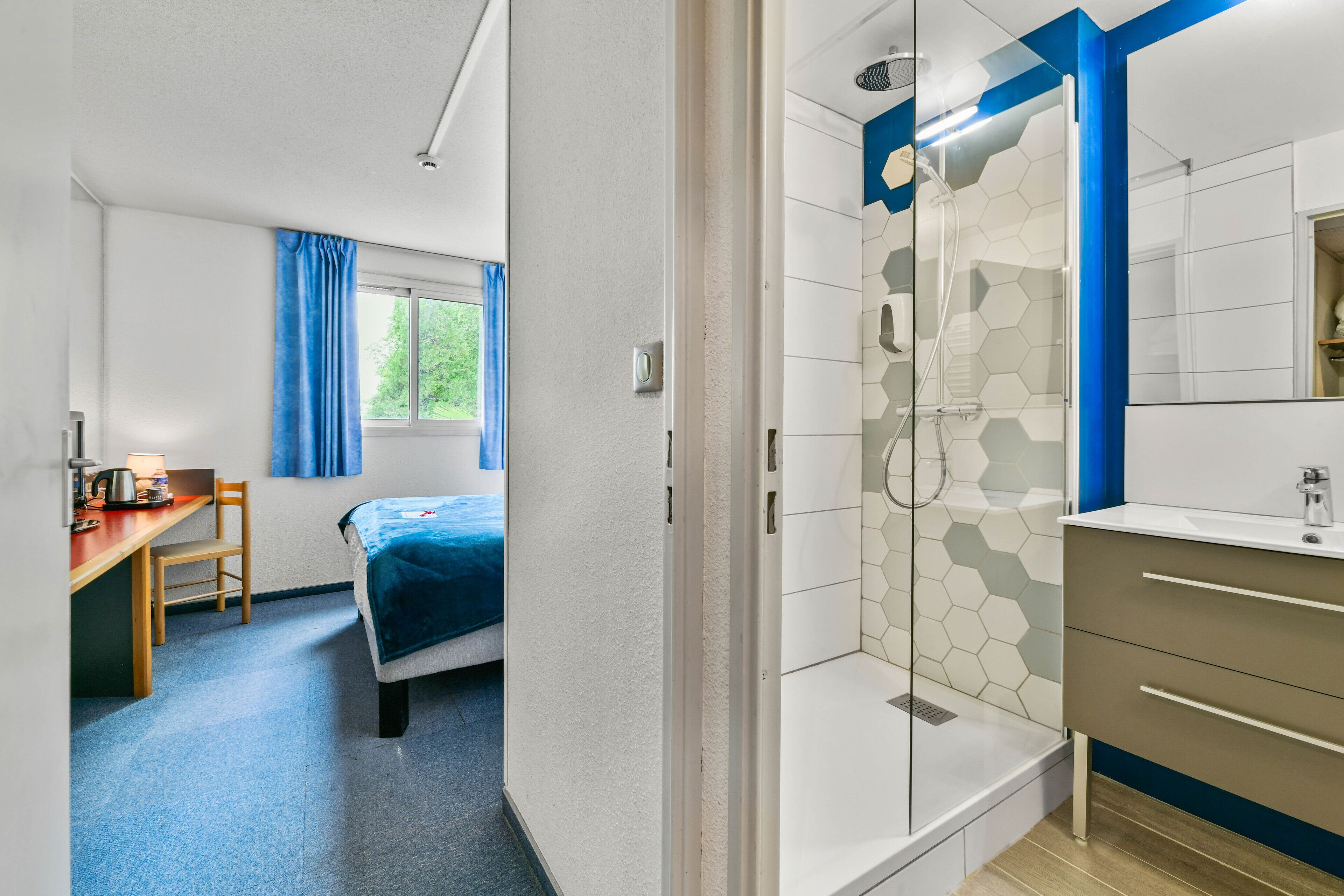 Een hotelkamer met een bed, een bureau, een douche en een badkamer met een spiegel en wastafel.