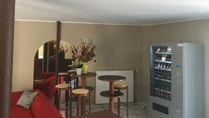 Un espace de vie compact comprenant un canapé rouge, une petite table à manger avec des tabourets, un miroir et un distributeur automatique.