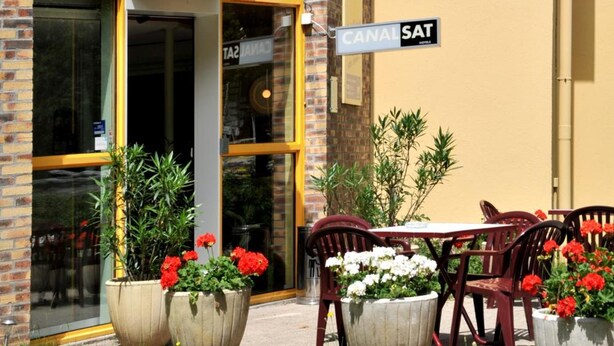 Un restaurant d’hôtel avec un auvent jaune, des tables en plein air décorées de fleurs rouges et blanches, et un panneau indiquant « CANAL SAT ».