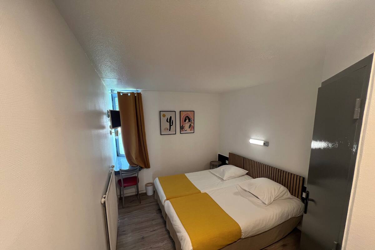 A hotel room with a bed, a desk, a chair, a window with curtains, and two wall-mounted pictures.