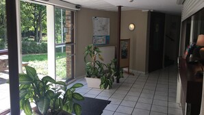 Un hall d’hôtel avec un sol carrelé, des plantes en pot, une carte accrochée au mur et une vue sur l’extérieur grâce à de grandes fenêtres.