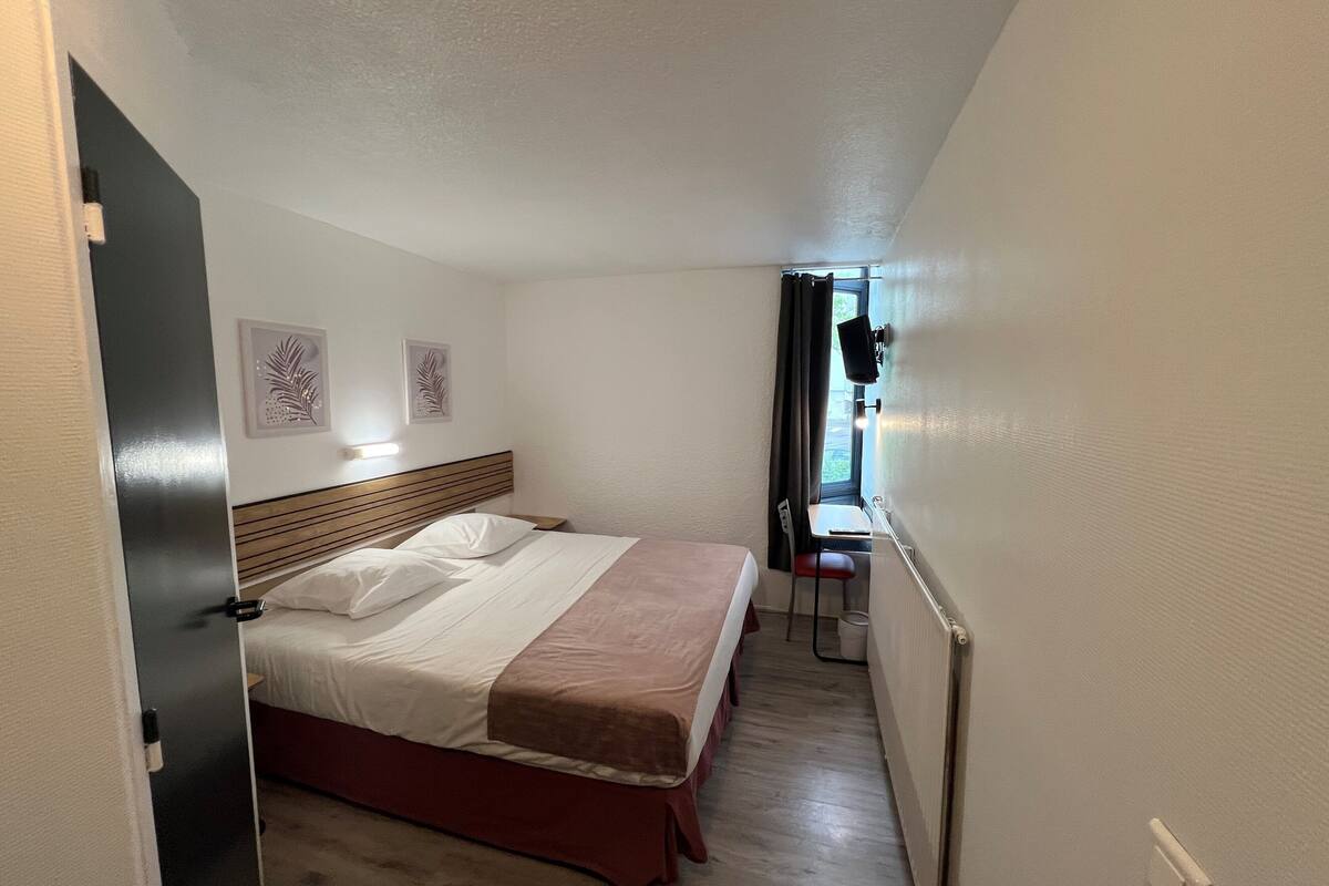 A narrow hotel room with a bed, a small table, and a window with curtains.