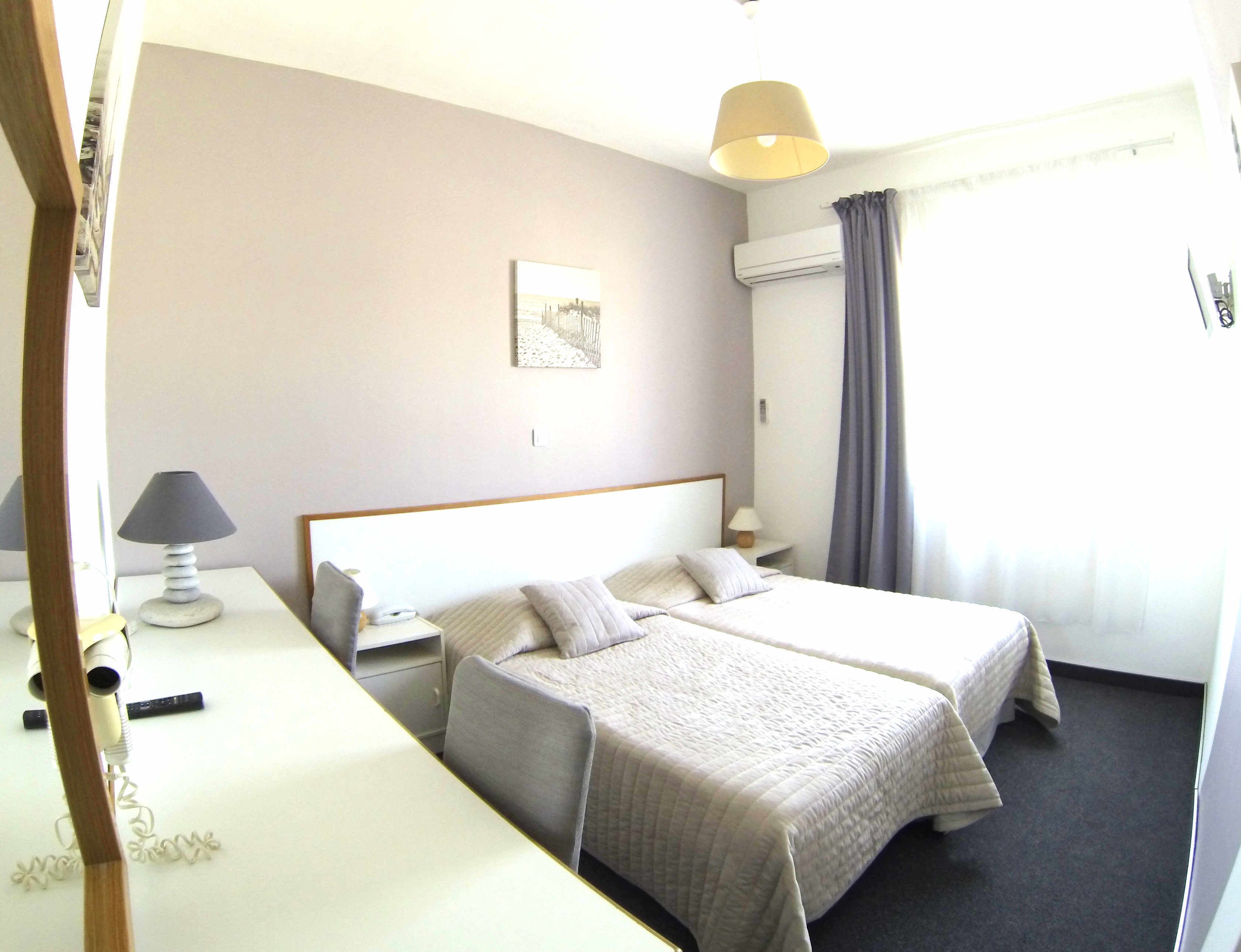 Habitación de hotel con dos camas, una mesita de noche con lámpara, otra mesita de noche más pequeña con lámpara, aire acondicionado de pared y una ventana con cortinas.