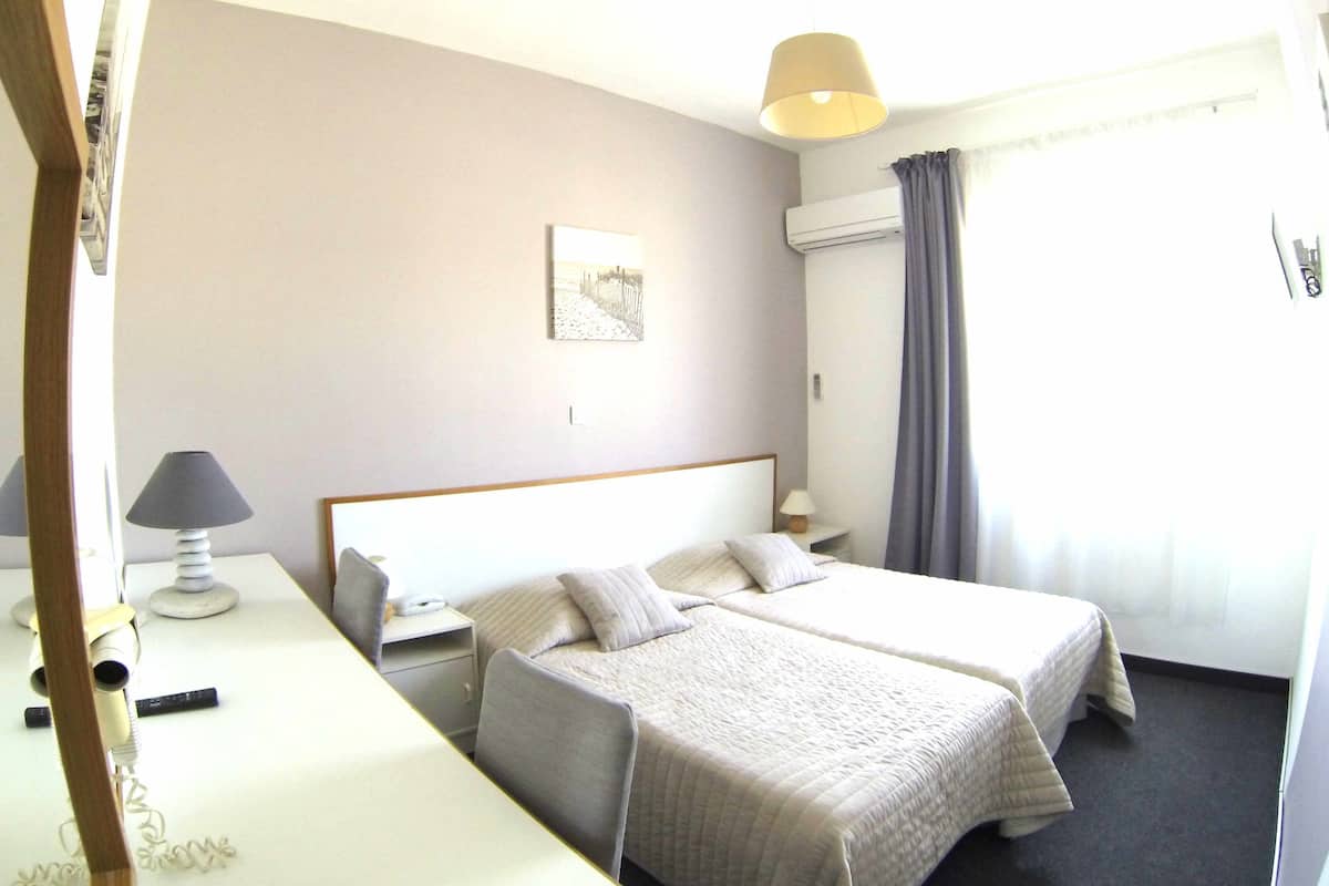 A hotel room with two beds, a bedside table with a lamp, a wall-mounted air conditioner, and a window with curtains.