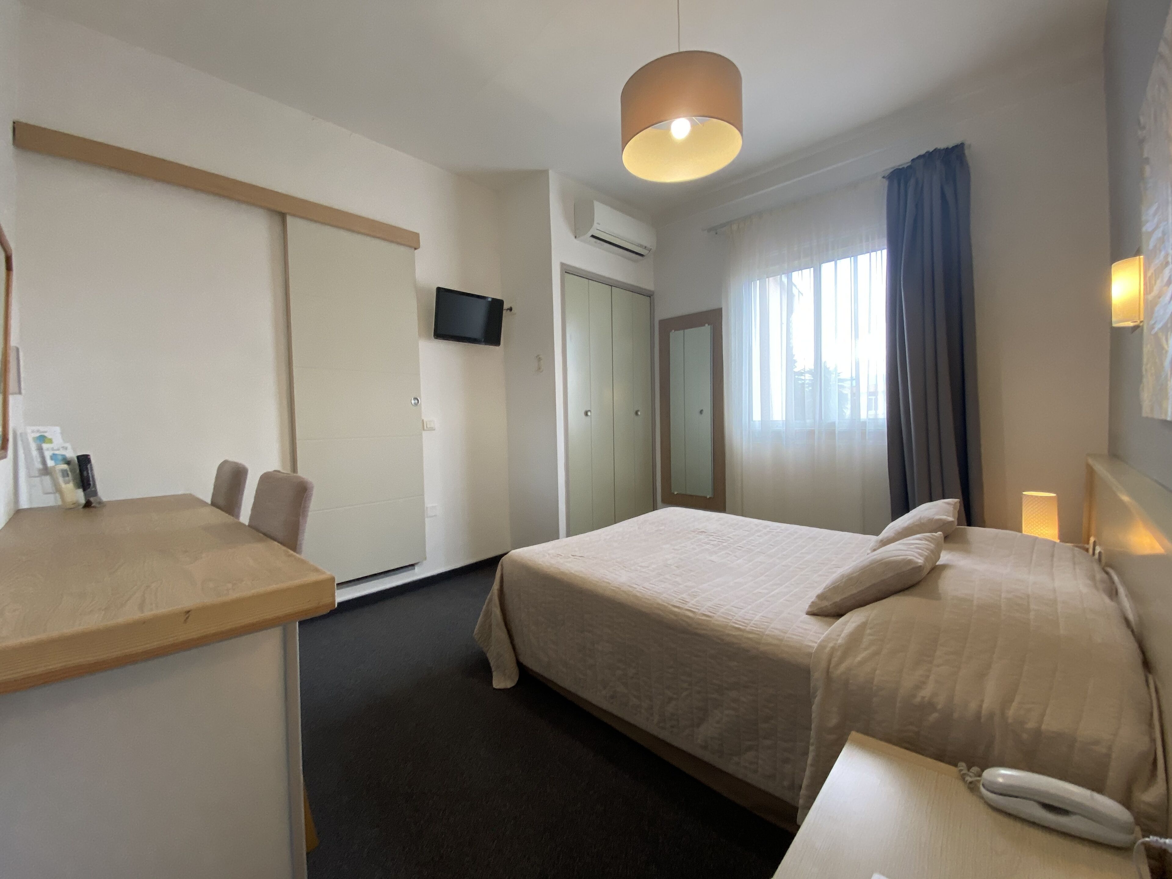 A hotel room with a bed, bedside table, and a desk with chairs. There is a TV mounted on the wall, a window with curtains, and a lamp on the bedside table.