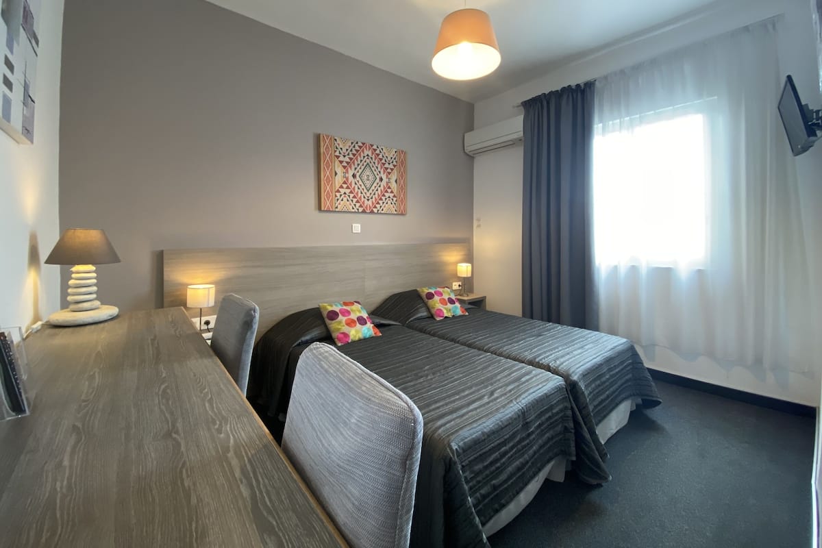 A hotel room with two beds, a desk, a lamp, a painting on the wall, and a window with curtains.