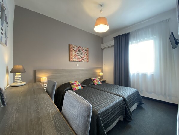 A hotel room with two beds, a desk, a lamp, a painting on the wall, and a window with curtains.