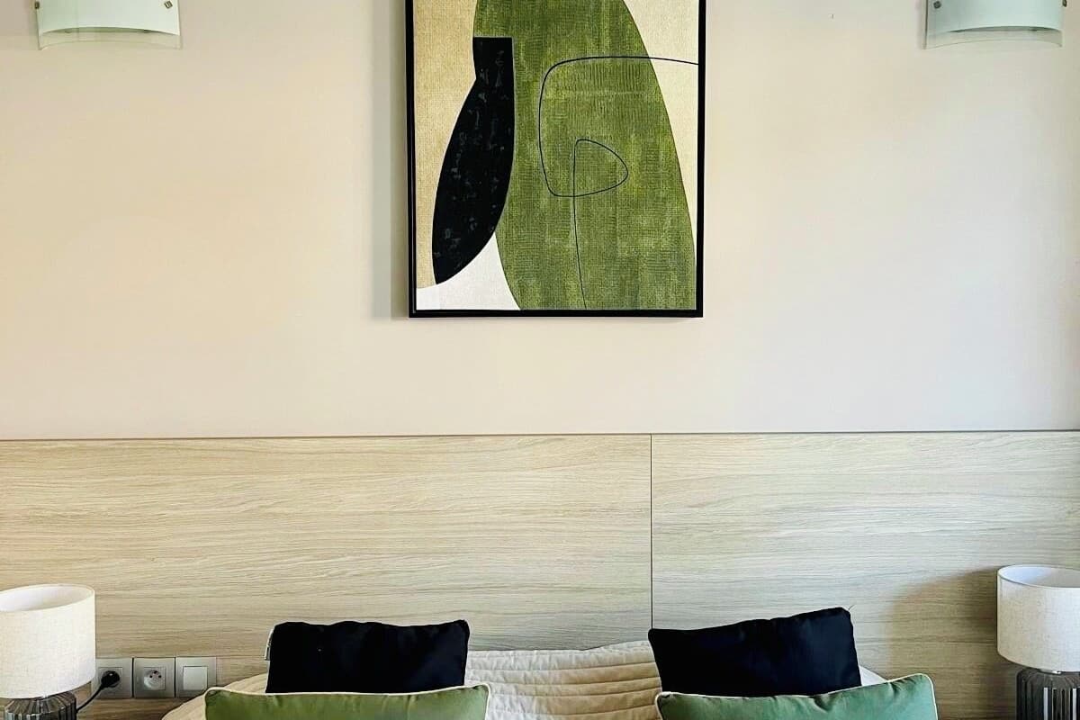 A hotel room with a bed, two green pillows, a white quilted bedspread, a wooden headboard, and a modern abstract painting on the wall.
