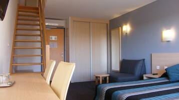 Camera d'albergo con una scrivania in legno, due letti con lenzuola blu, un piccolo tavolo rotondo, uno specchio e una scala in legno che conduce a un armadio.