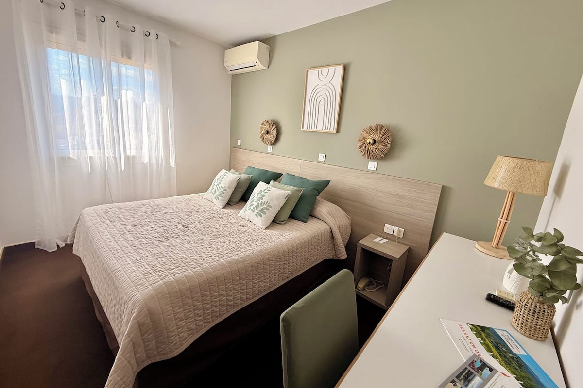 A hotel room with a bed, a desk, a chair, an air conditioner, and a lamp.