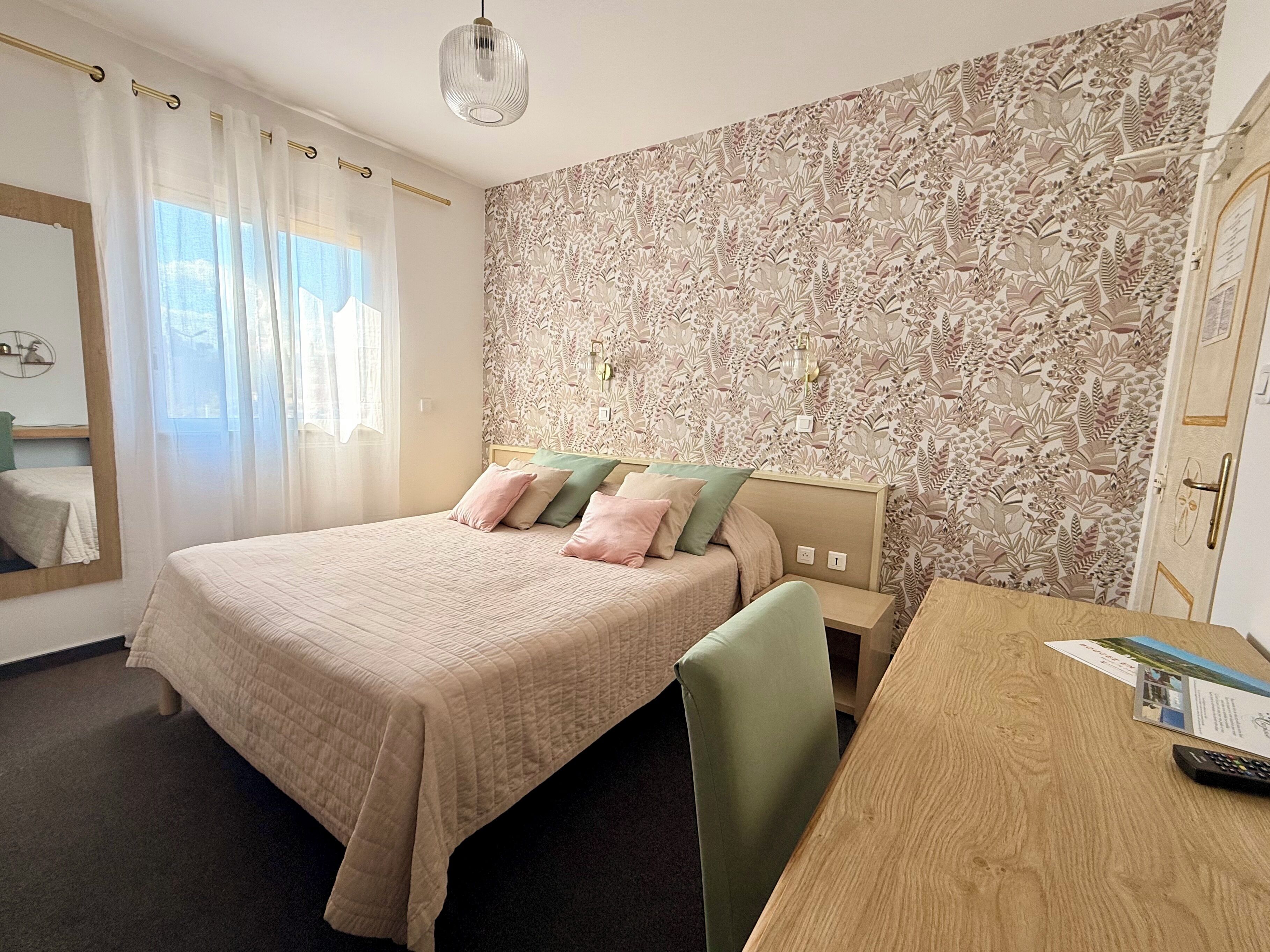 A bedroom with a bed, a desk, a chair, a mirror, and a patterned wall.