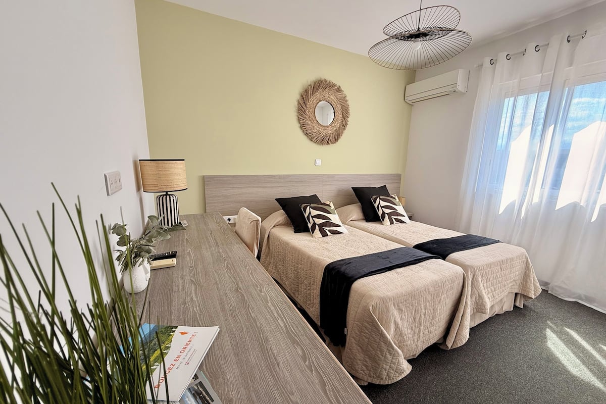 A hotel room with two beds, a bedside table, a lamp, a mirror, a wall decoration, and a window with curtains.