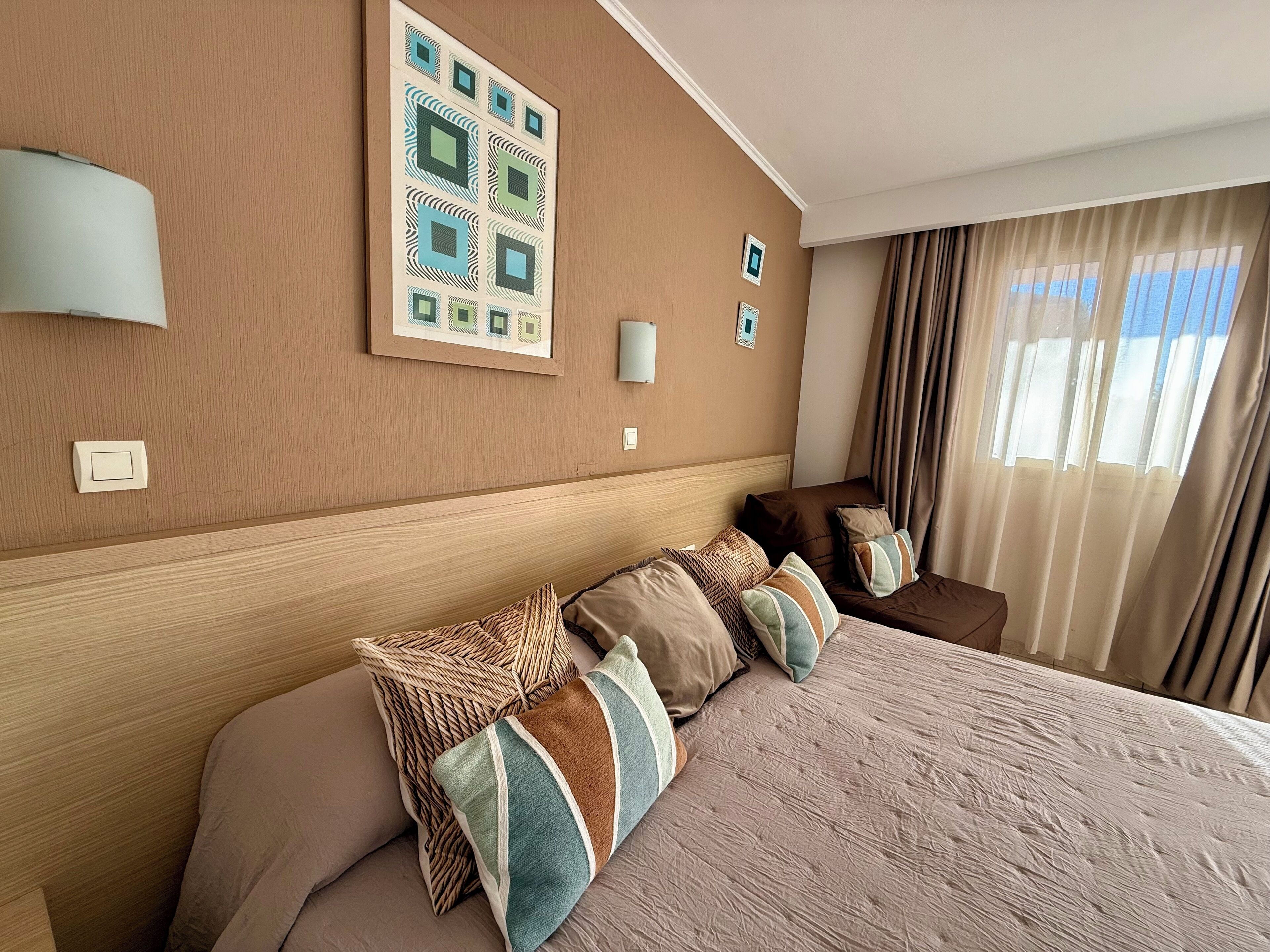 Habitación de hotel con una cama grande, varios almohadones, cabecera, decoración en la pared y una ventana con cortinas.