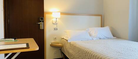Habitación de hotel con cama, mesita de noche, lámpara, teléfono y puerta.