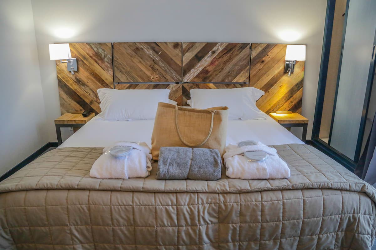 Habitación de hotel con cabecera de madera, dos mesitas de noche con lámparas, cama con colcha color beige y toallas dispuestas sobre la cama.