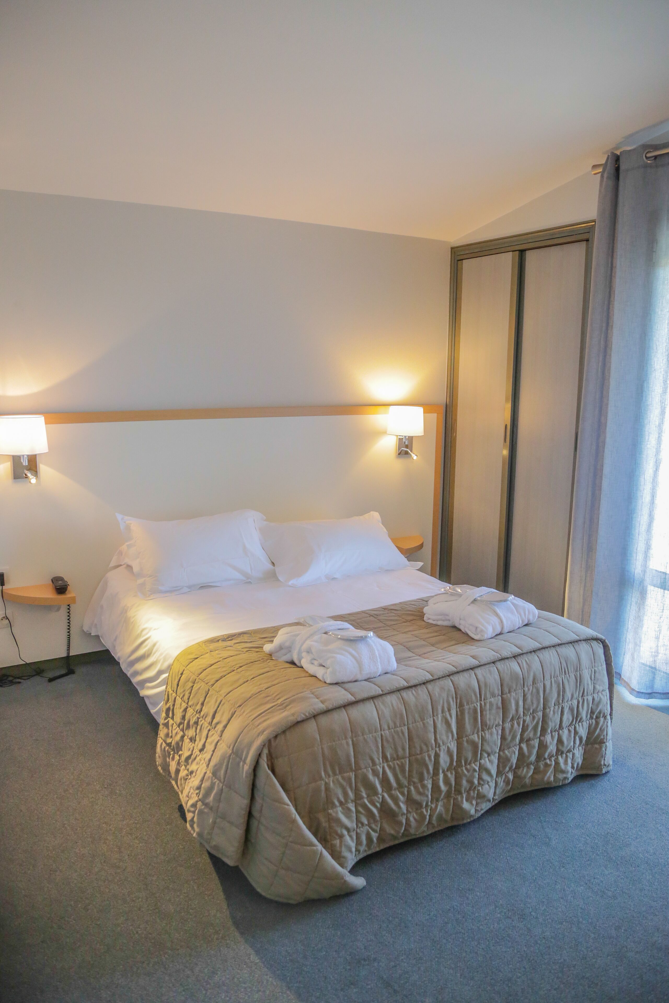 A hotel room with a bed, two bedside lamps, a nightstand, and a large window with curtains.