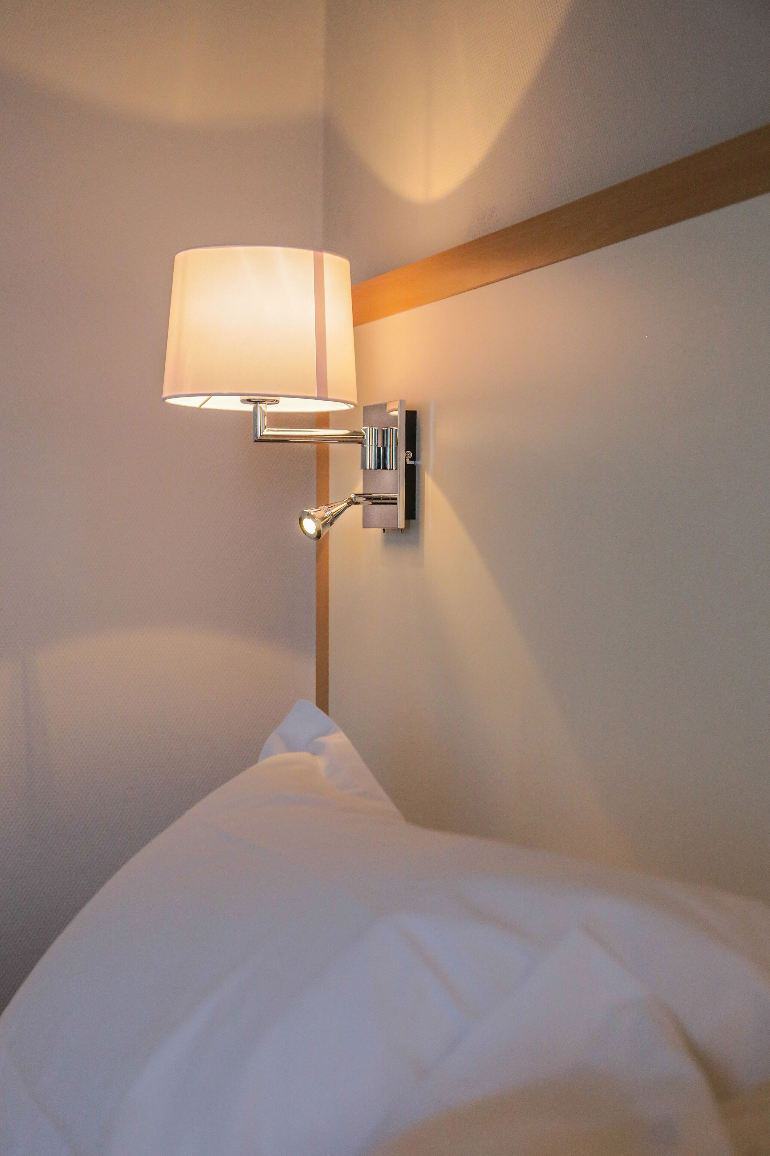 Una cama bien hecha con edredón blanco y almohadas, una lámpara de pared con pantalla blanca, y un cabecero de madera.