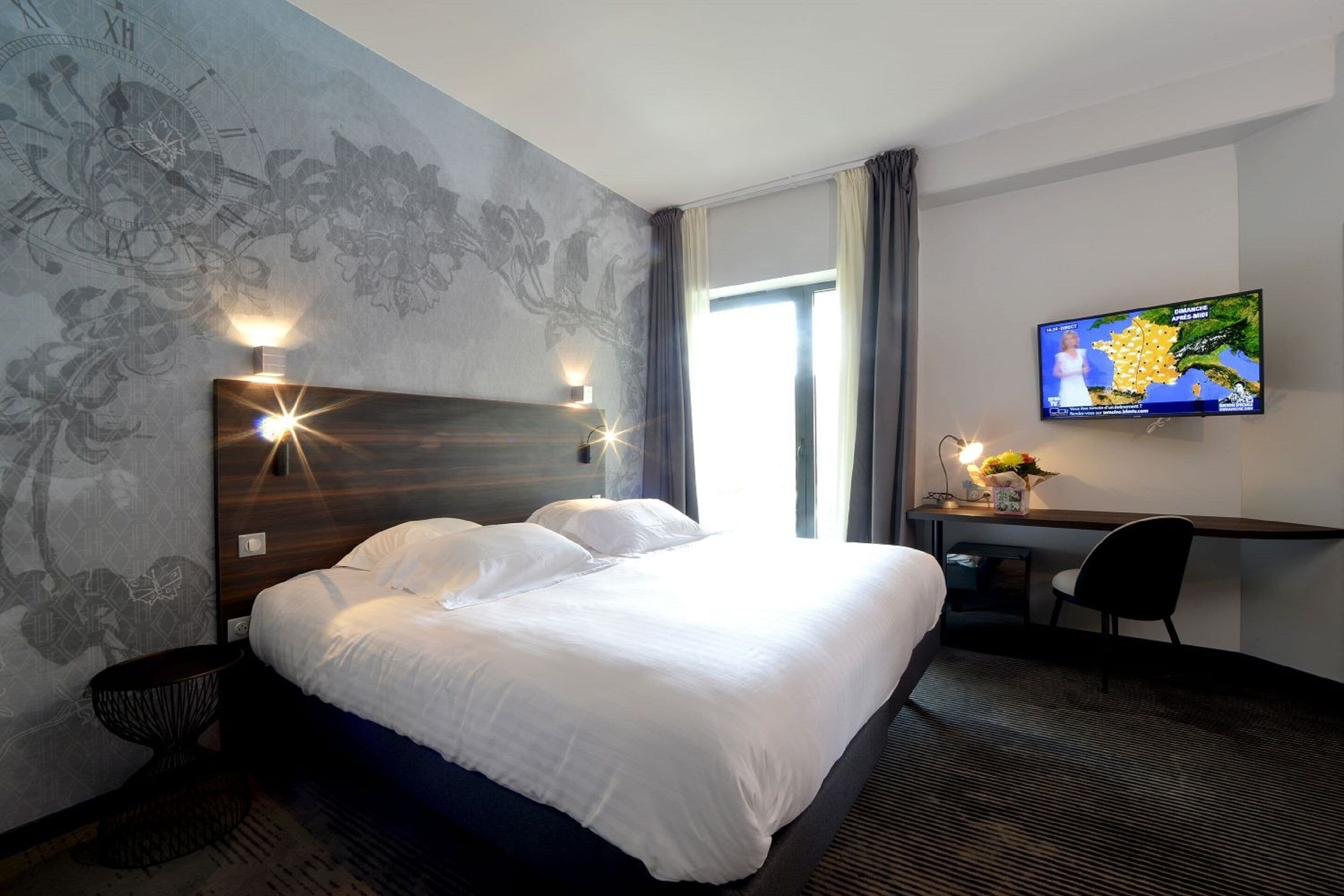 A hotel room with a large bed, a desk with a chair, a television mounted on the wall, and a window with curtains.