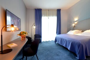 Una habitación de hotel con cama, escritorio, silla, lámpara y un cuadro en la pared.
