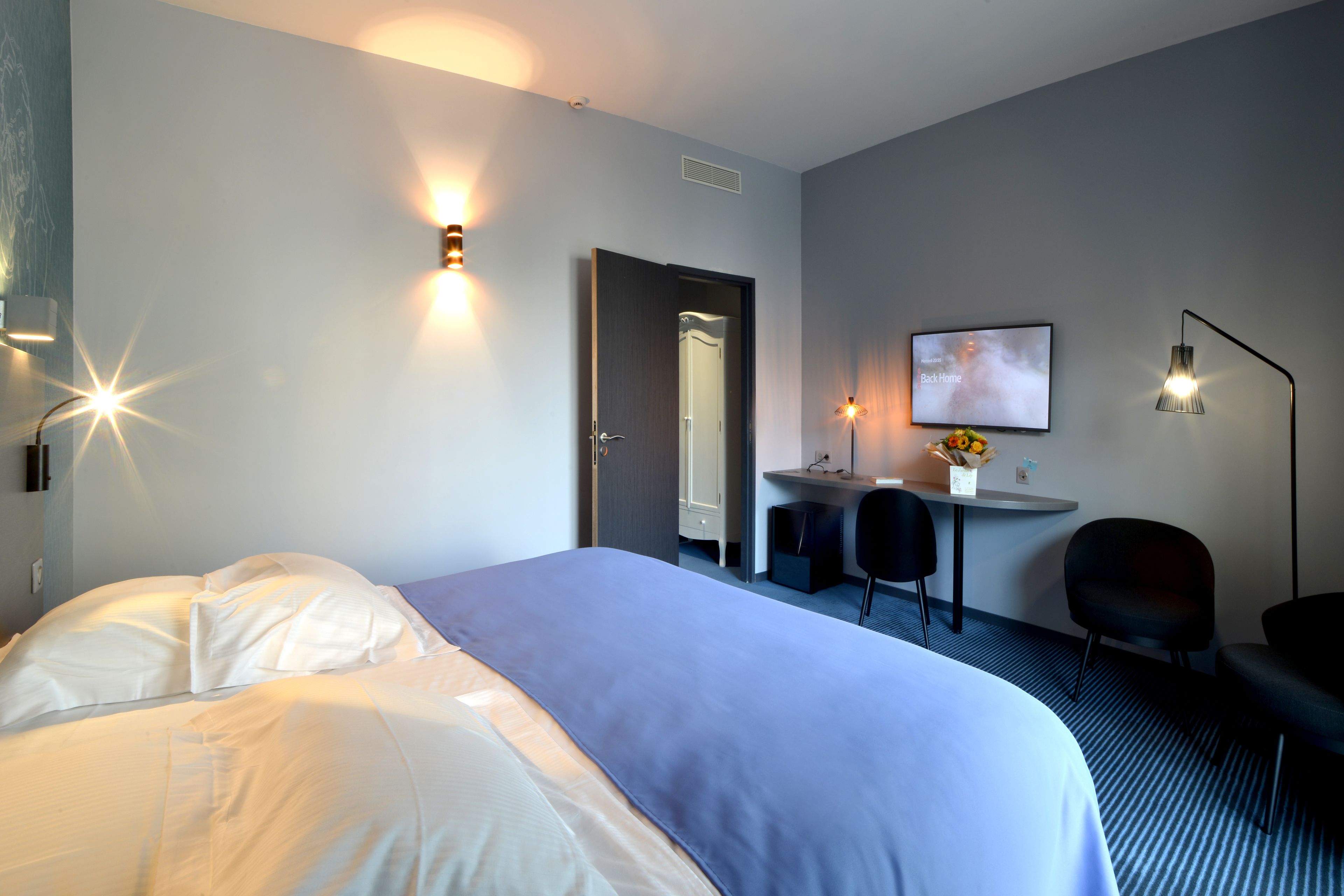 Habitación de hotel con una cama grande, un escritorio con silla, una mesita con un florero y un televisor montado en la pared.