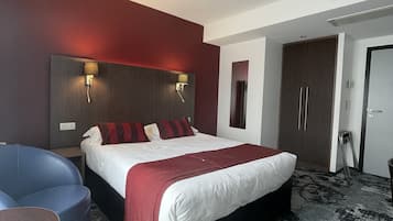 A hotel room with a large bed, a desk, and a chair. There is a wardrobe and a door visible in the background.