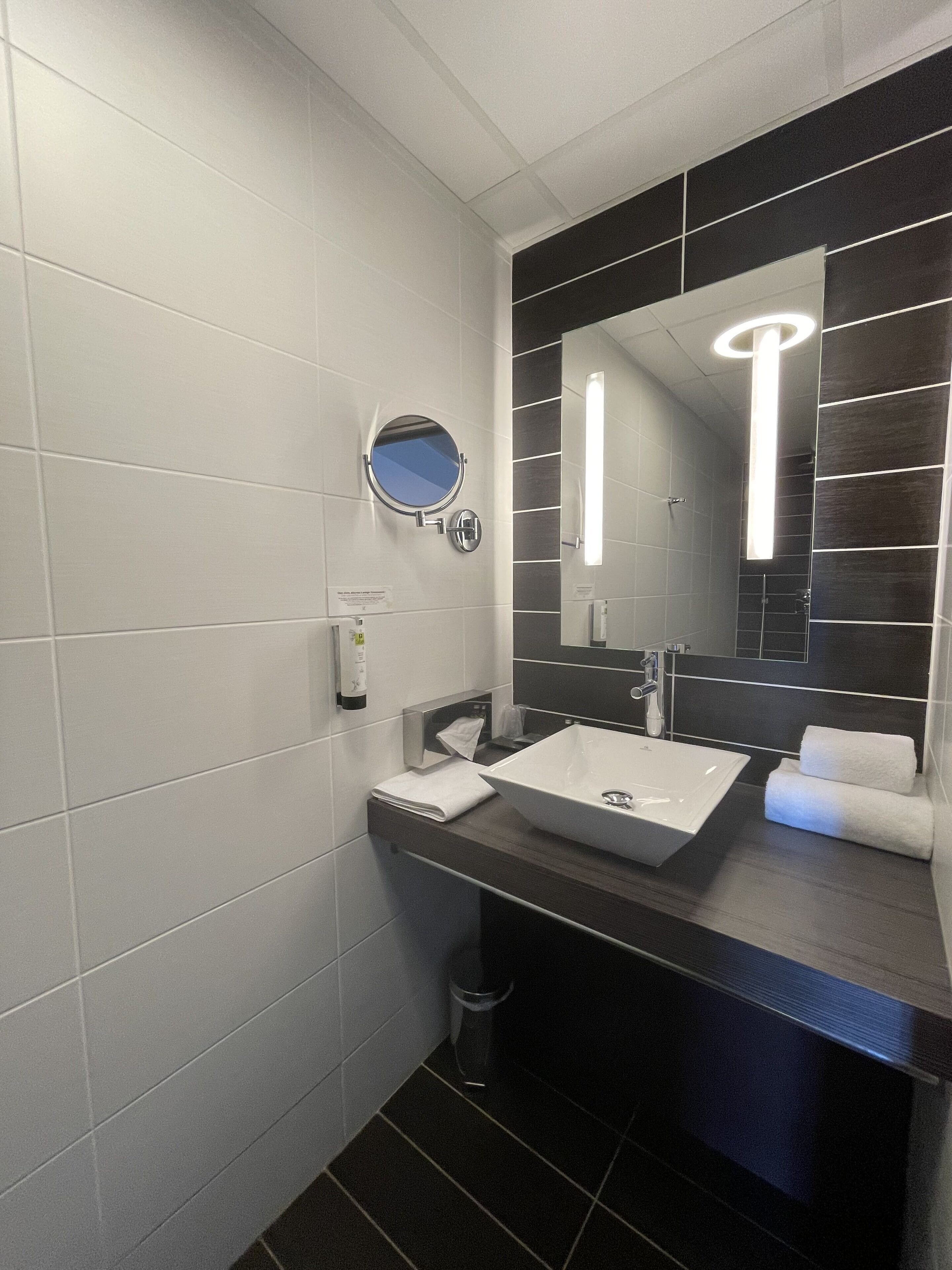 Un baño moderno con un lavamanos blanco, encimera oscura y un espejo grande con marco circular.