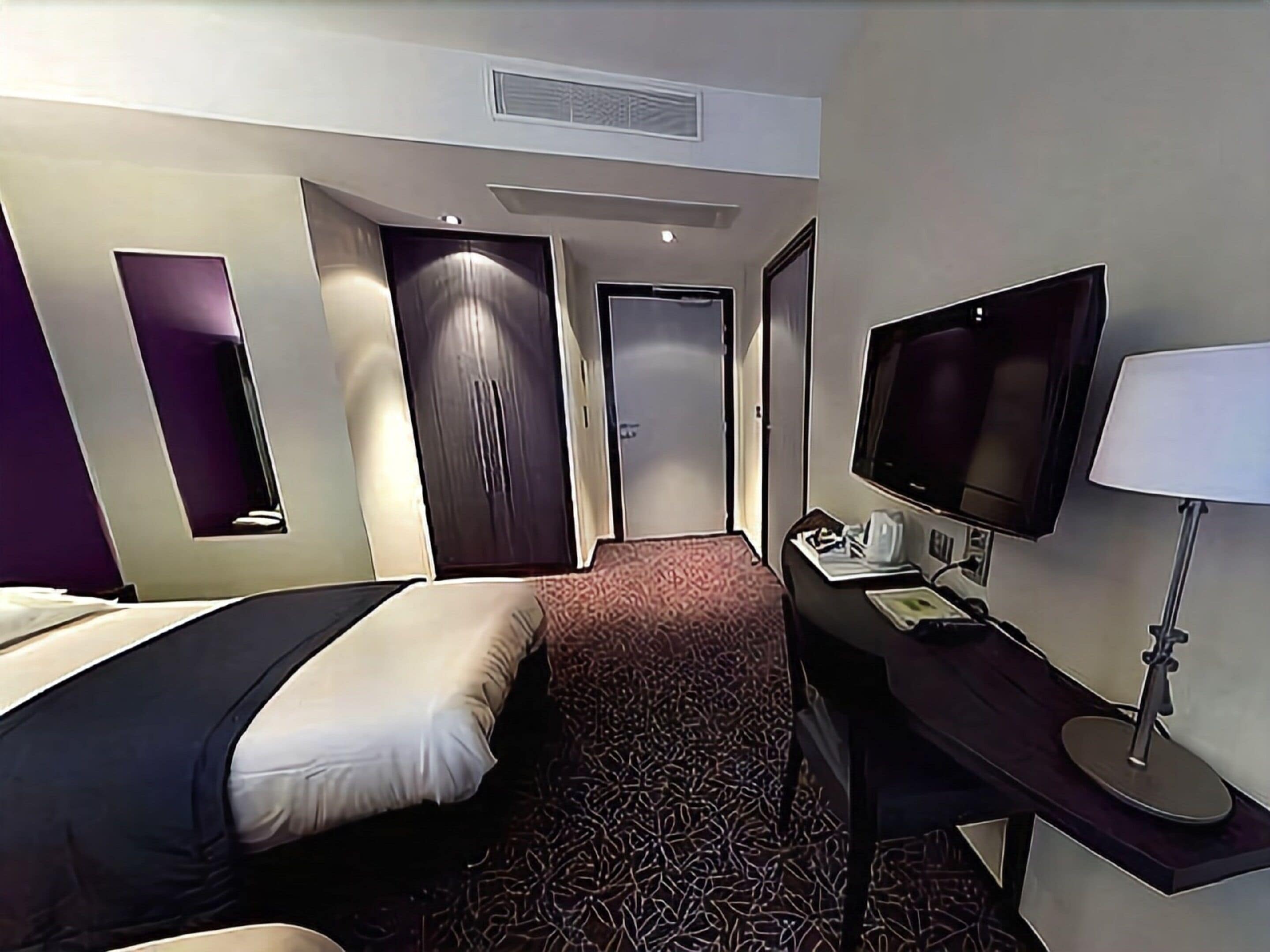 A hotel room with two beds, a desk with a computer and lamp, a TV, and a patterned carpet.