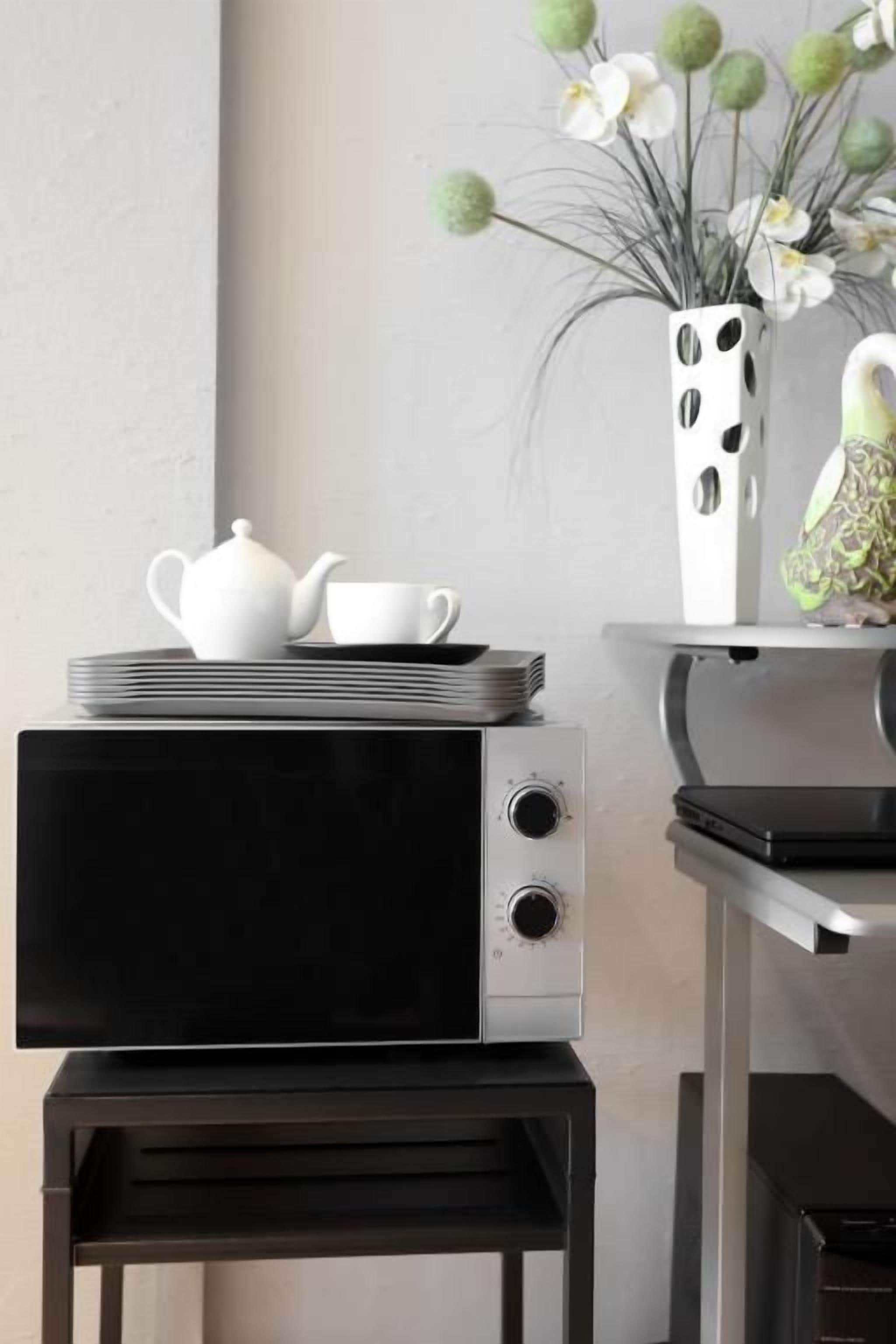 Un four à micro-ondes posé sur un support noir, une théière blanche et des tasses empilées sur une pile d’assiettes, et un vase blanc orné de décorations sphériques vertes, le tout sur une table.