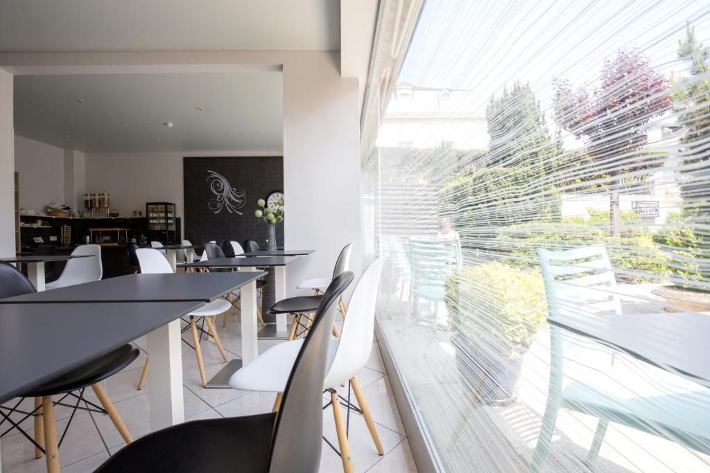 A modern dining area with a long table and chairs, a black wall with a decorative piece, and a view of greenery through a window with blinds.