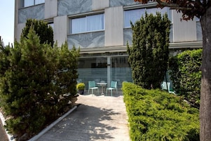 Un edificio con fachada moderna, un espacio al aire libre con pavimento y zona de estar, y abundante vegetación.