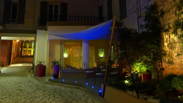 Un patio con suelo de adoquines, una zona de estar cubierta con sillas y mesas, y un edificio con varias ventanas y puertas.