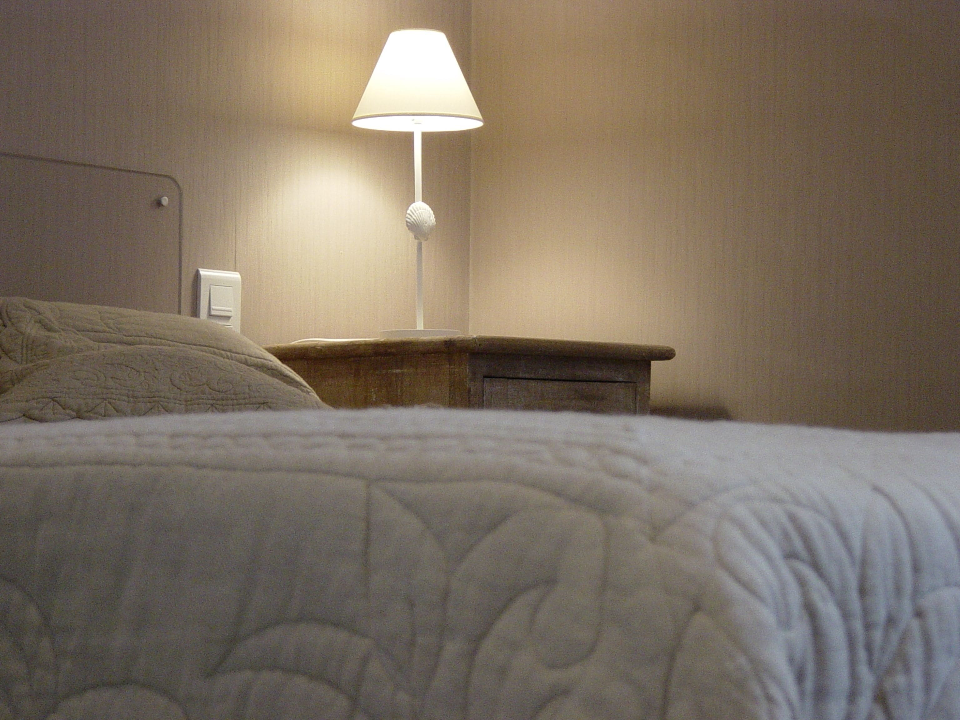 A neatly made bed with a white quilt, a bedside table with a lamp, and a wall socket.