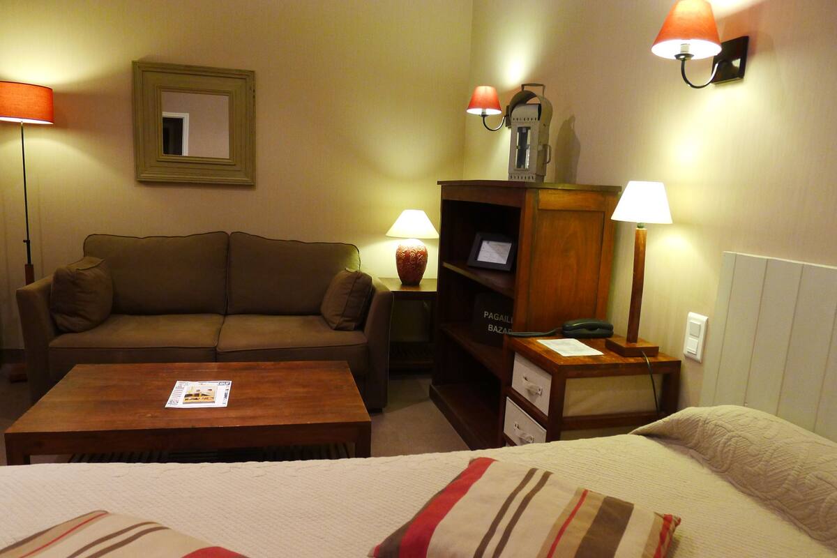 A hotel room with a sofa, a wooden table, a desk with a lamp, a bookshelf, and a bed with striped pillows.