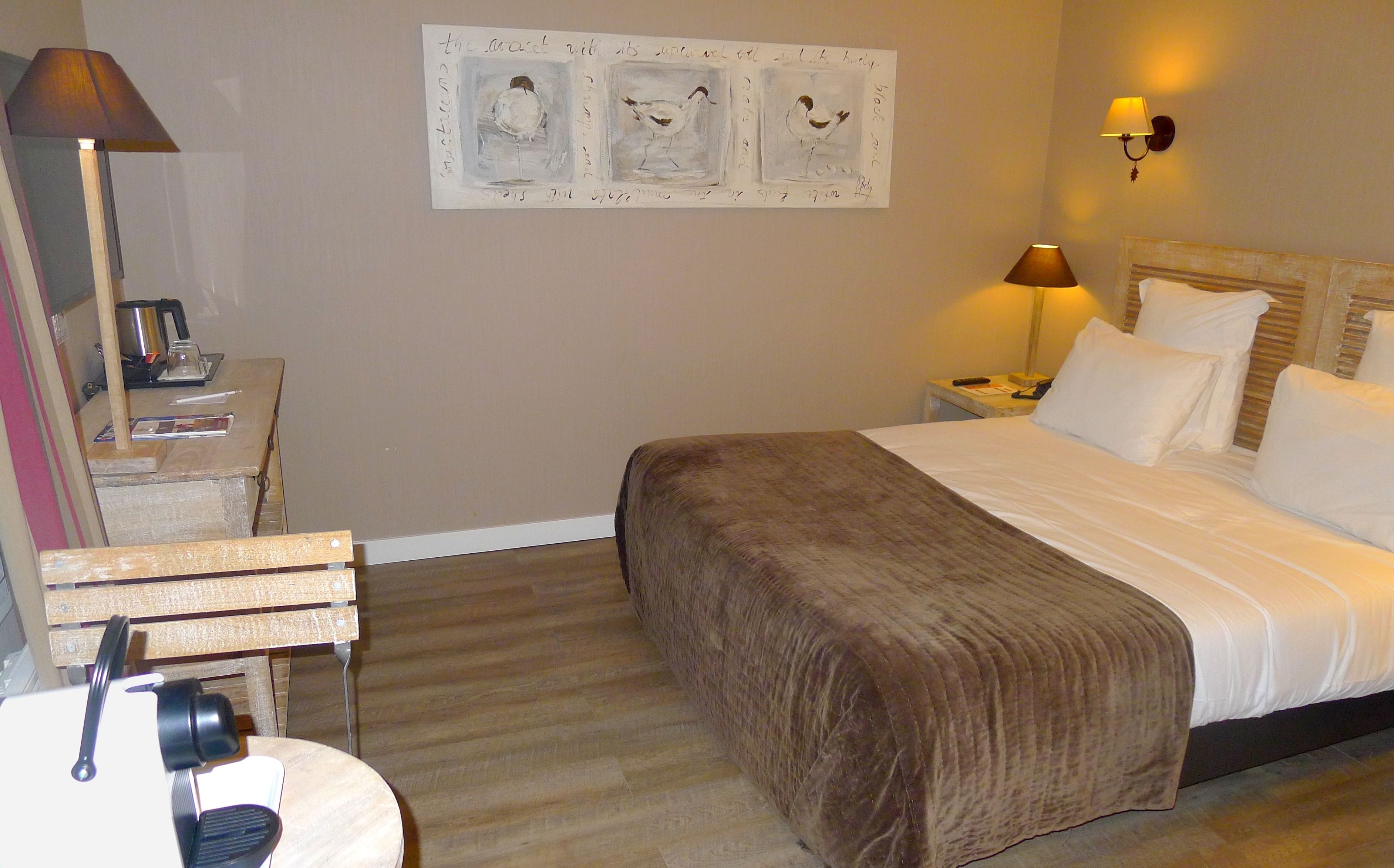 Habitación de hotel con cama, escritorio con lámpara, una pared decorada con obras de arte y mesita de noche con lámpara.