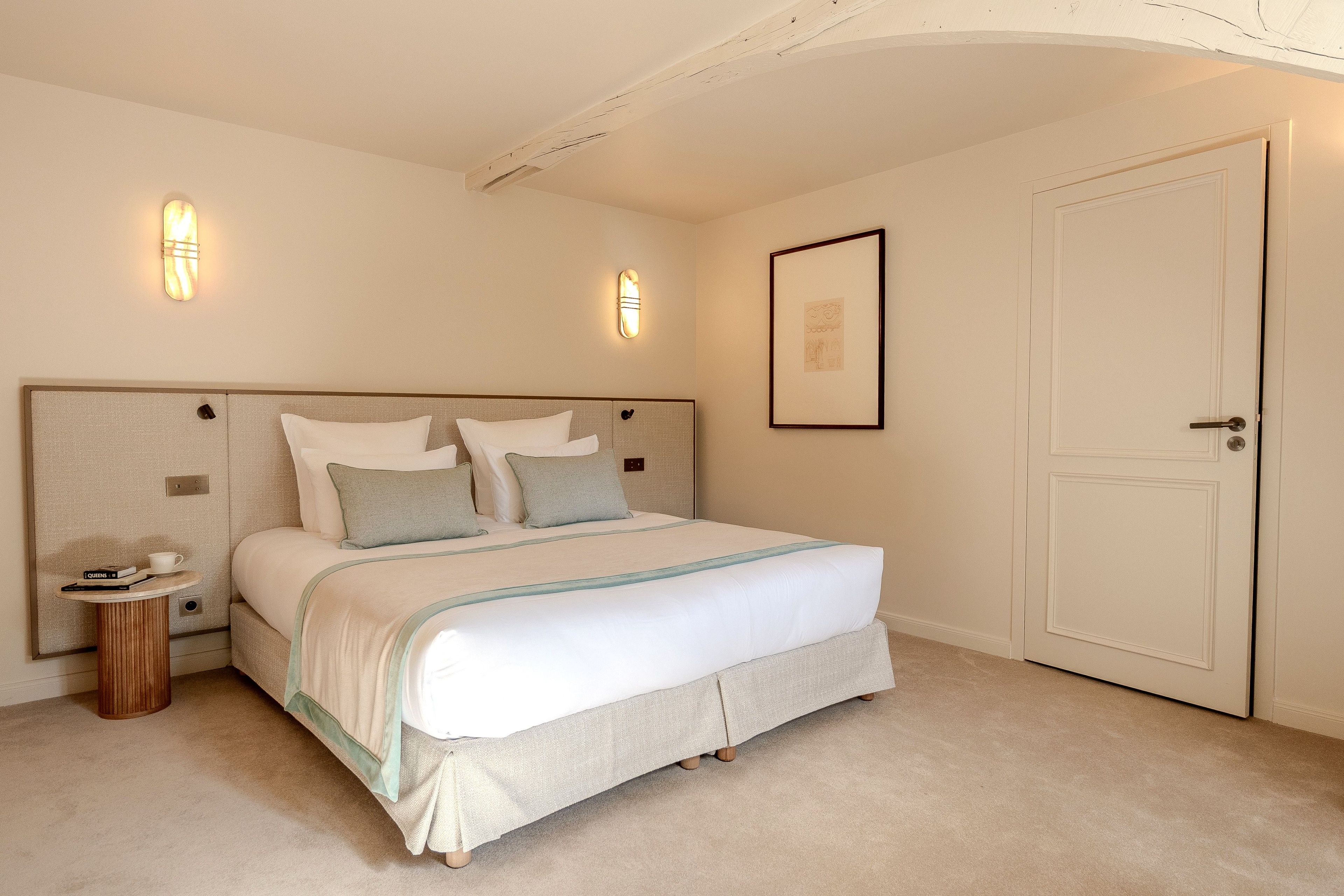 Un dormitorio con cama, mesita de noche, luces empotradas en la pared, un cuadro enmarco y una puerta.