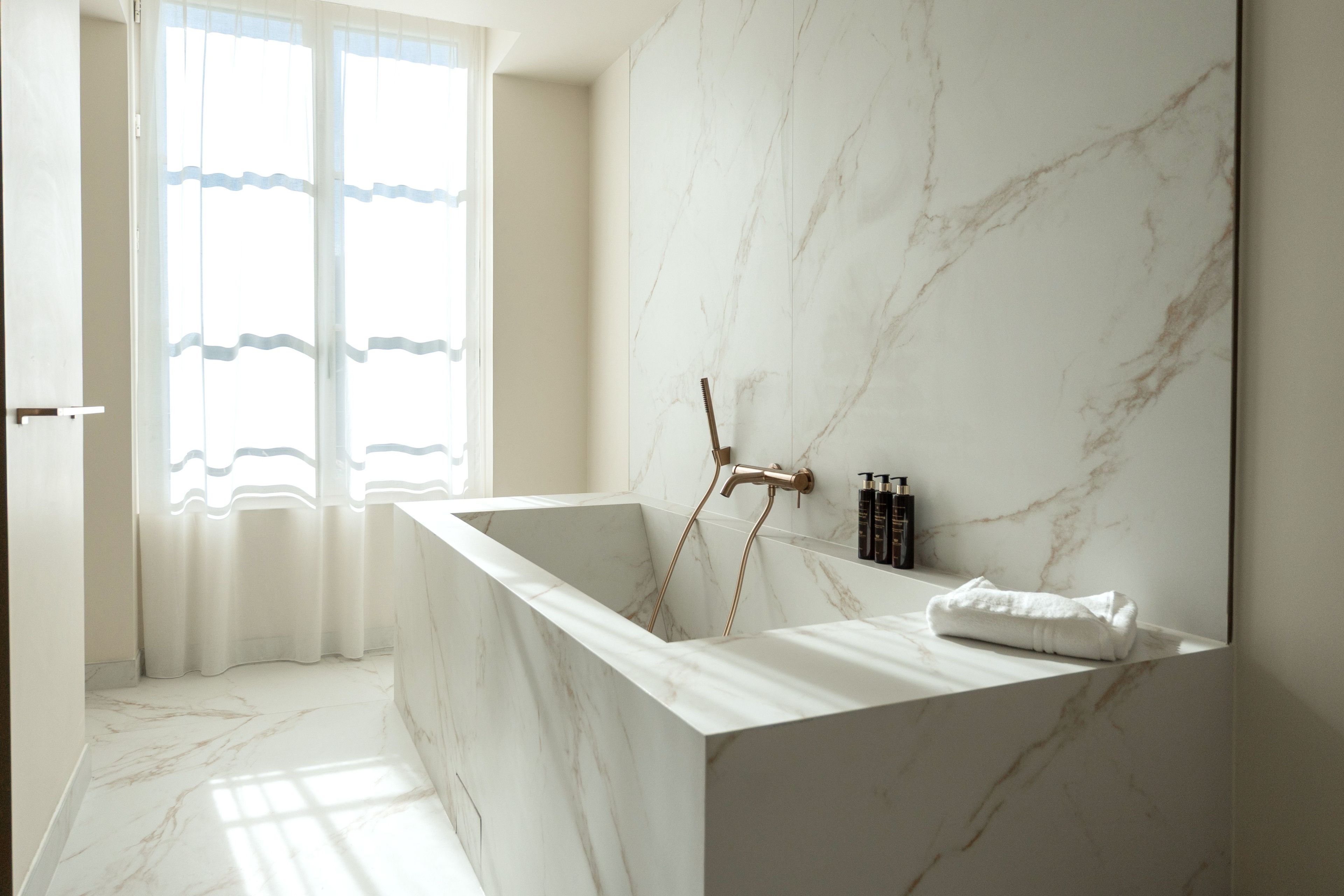 A modern bathroom with a marble sink, a shower with a rain showerhead, and a large window with sheer curtains.