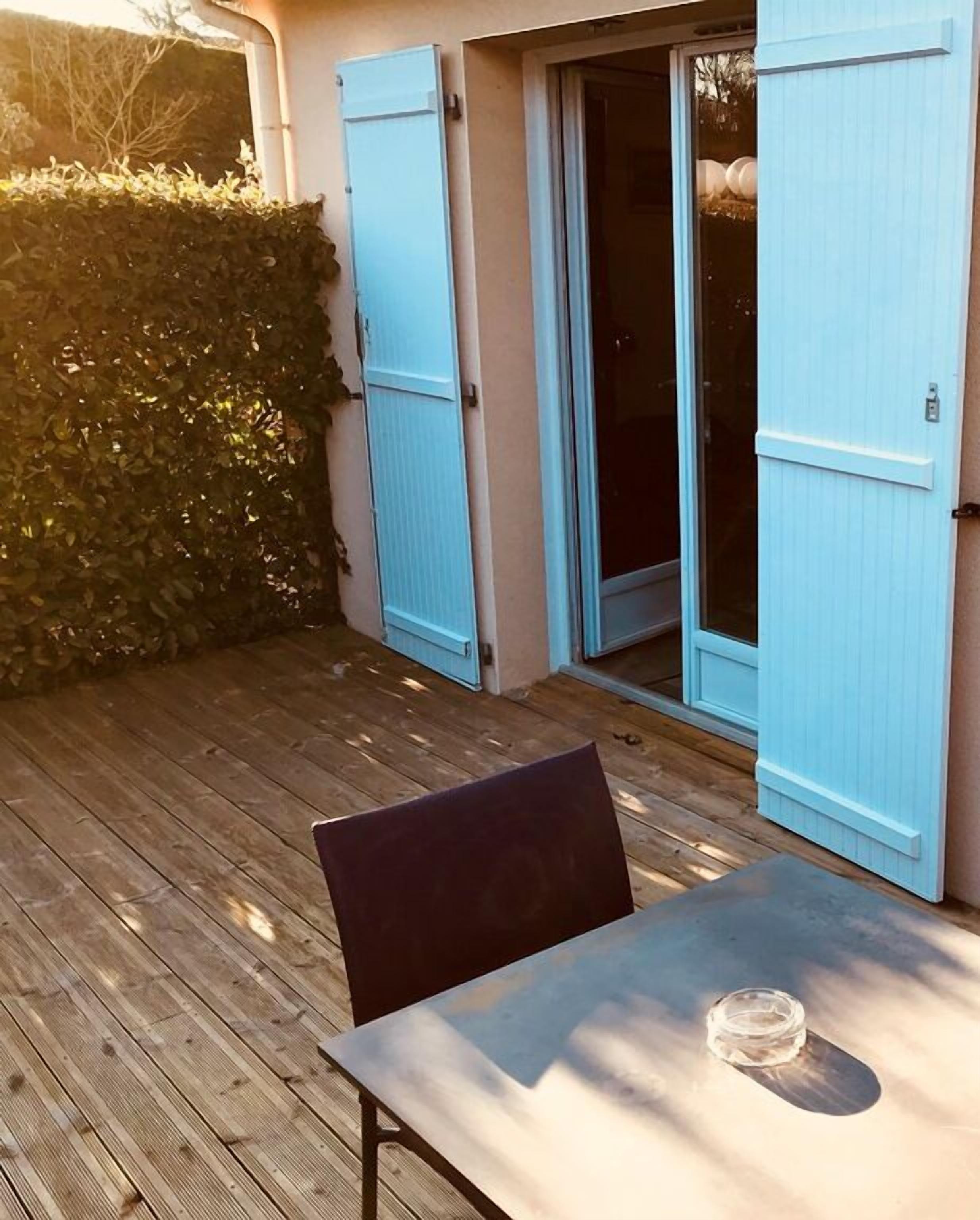 A wooden deck with a table and chairs, a blue door, and a hedge in the background.