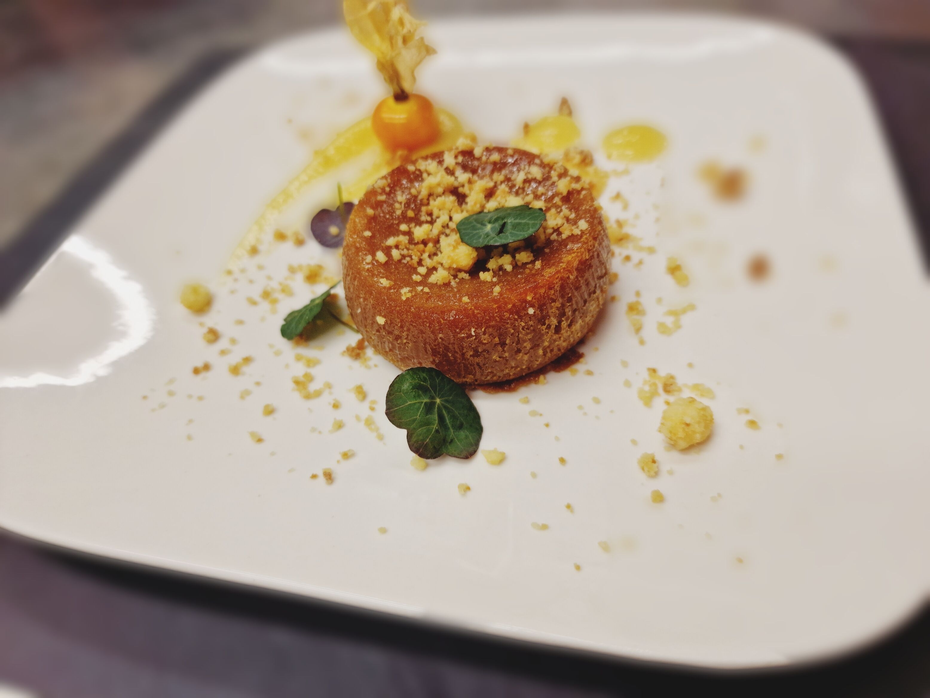 Een vierkant bord met een rond, goudbruin gerecht, versierd met groene bladeren en gele kruimels.
