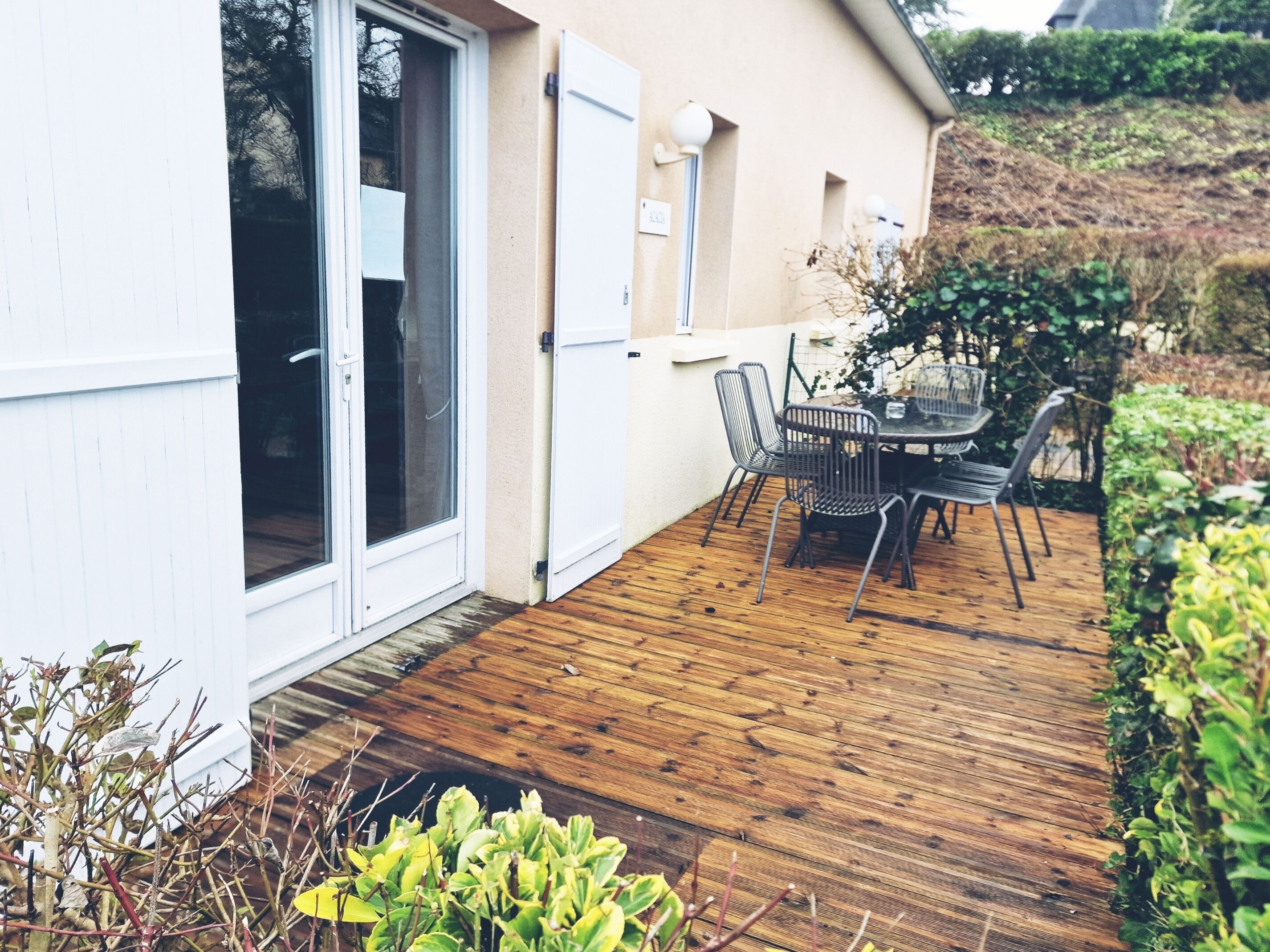 Terraza de madera con mesa y sillas, una puerta corrediza de vidrio y una pared exterior blanca.