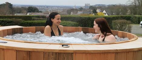 Ein hölzernes Whirlpool mit zwei Personen darin, umgeben von einem Garten und mit Blick auf eine Stadt im Hintergrund.