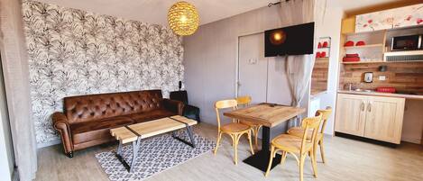 A compact living space with a brown leather sofa, a wooden dining table with four chairs, a patterned wall, a ceiling light, and a kitchen area with wooden cabinets and a microwave.